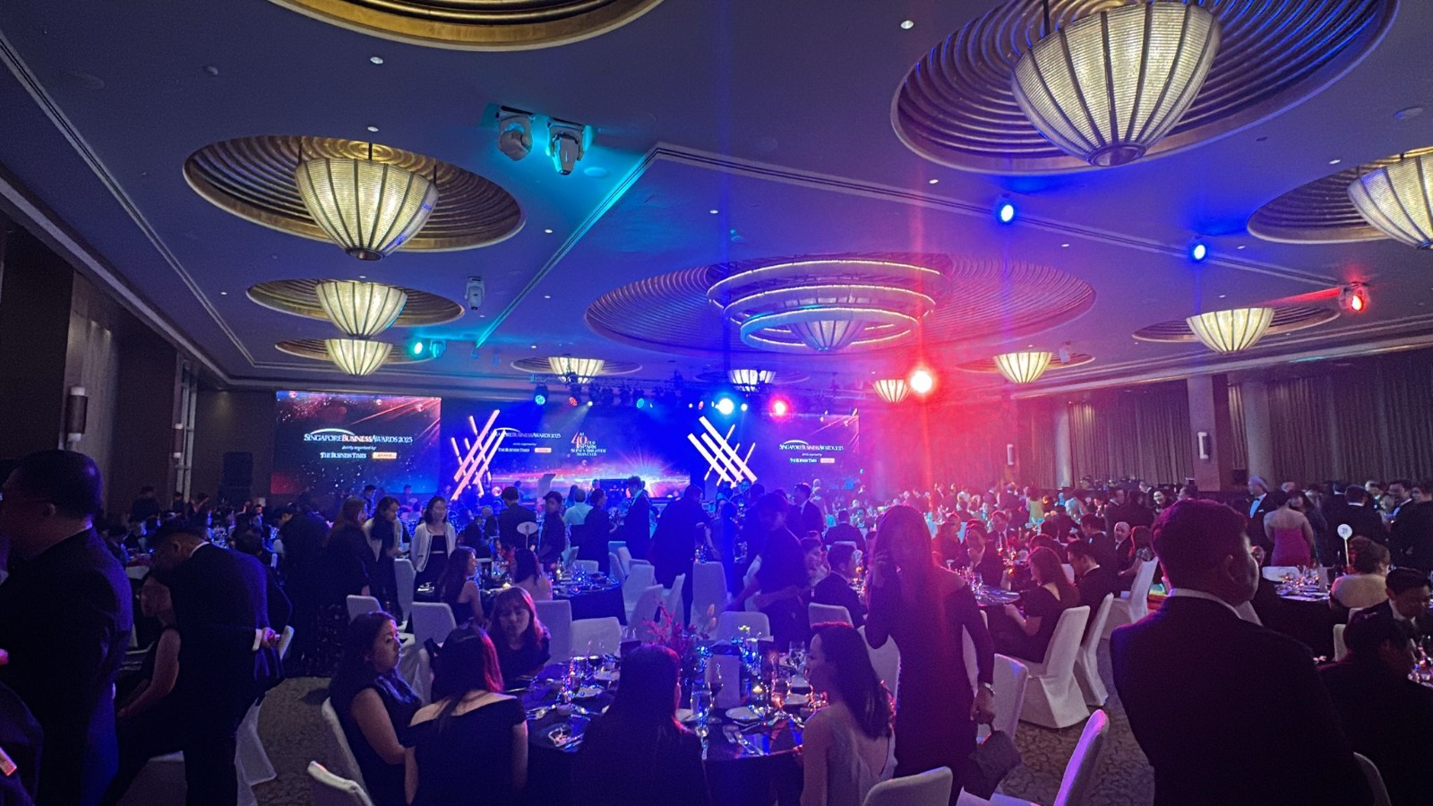 Singapore Business Awards 2025 gala dinner — packed ballroom with RGB stage lighting, round banquet tables, black-tie guests and chandelier ceiling